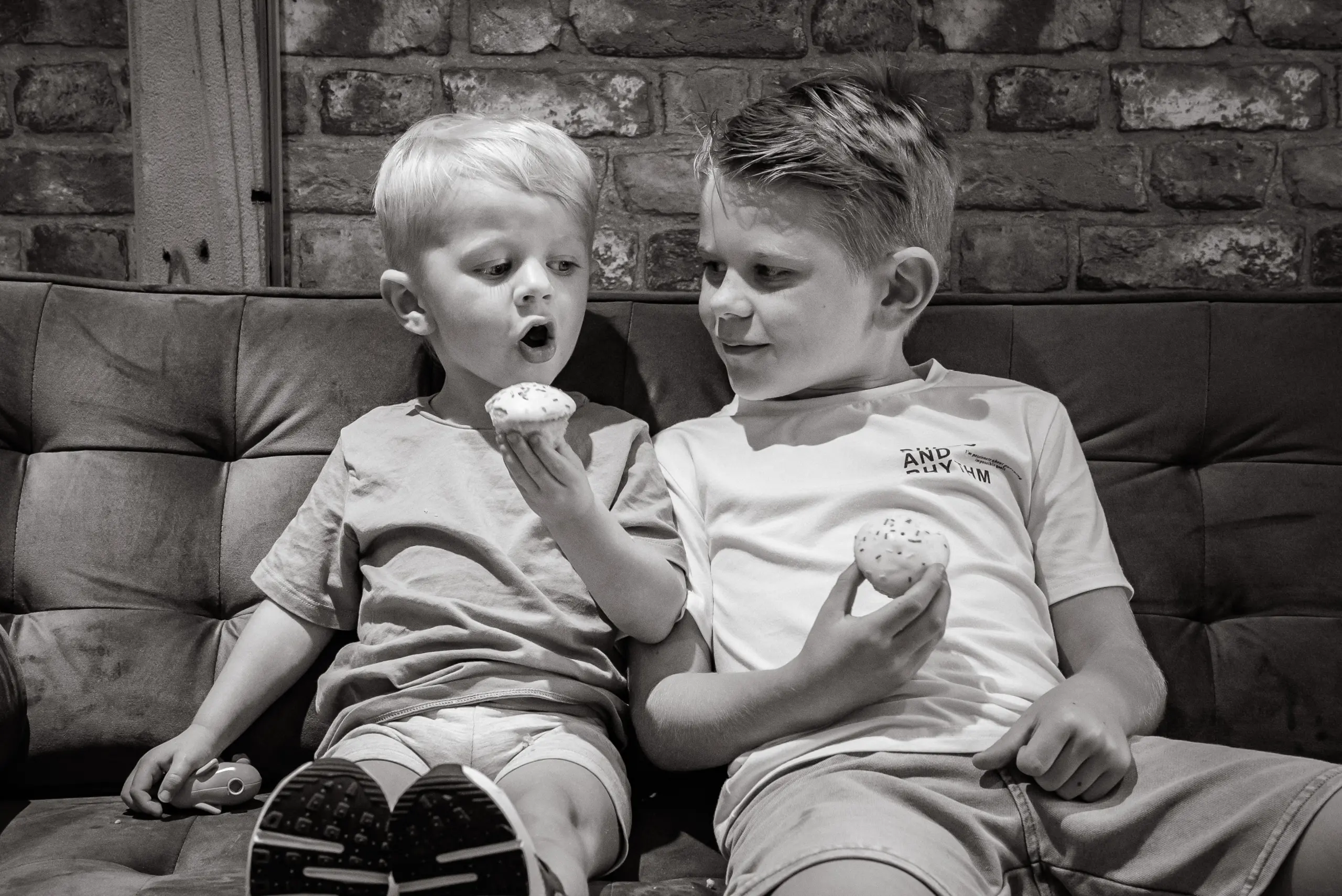 Two young children sitting on a sofa eating cupcakes, with a brick wall behind them