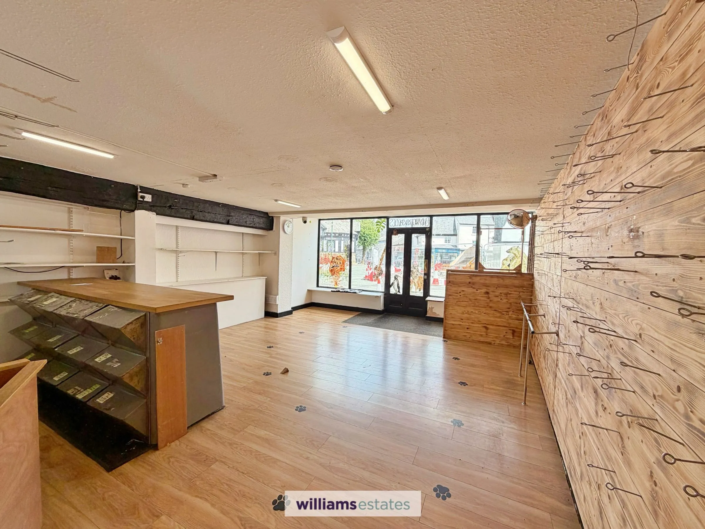 Empty retail shop interior with wooden flooring, display counter on left, white shelving, brick wall on right with metal hooks, and storefront windows