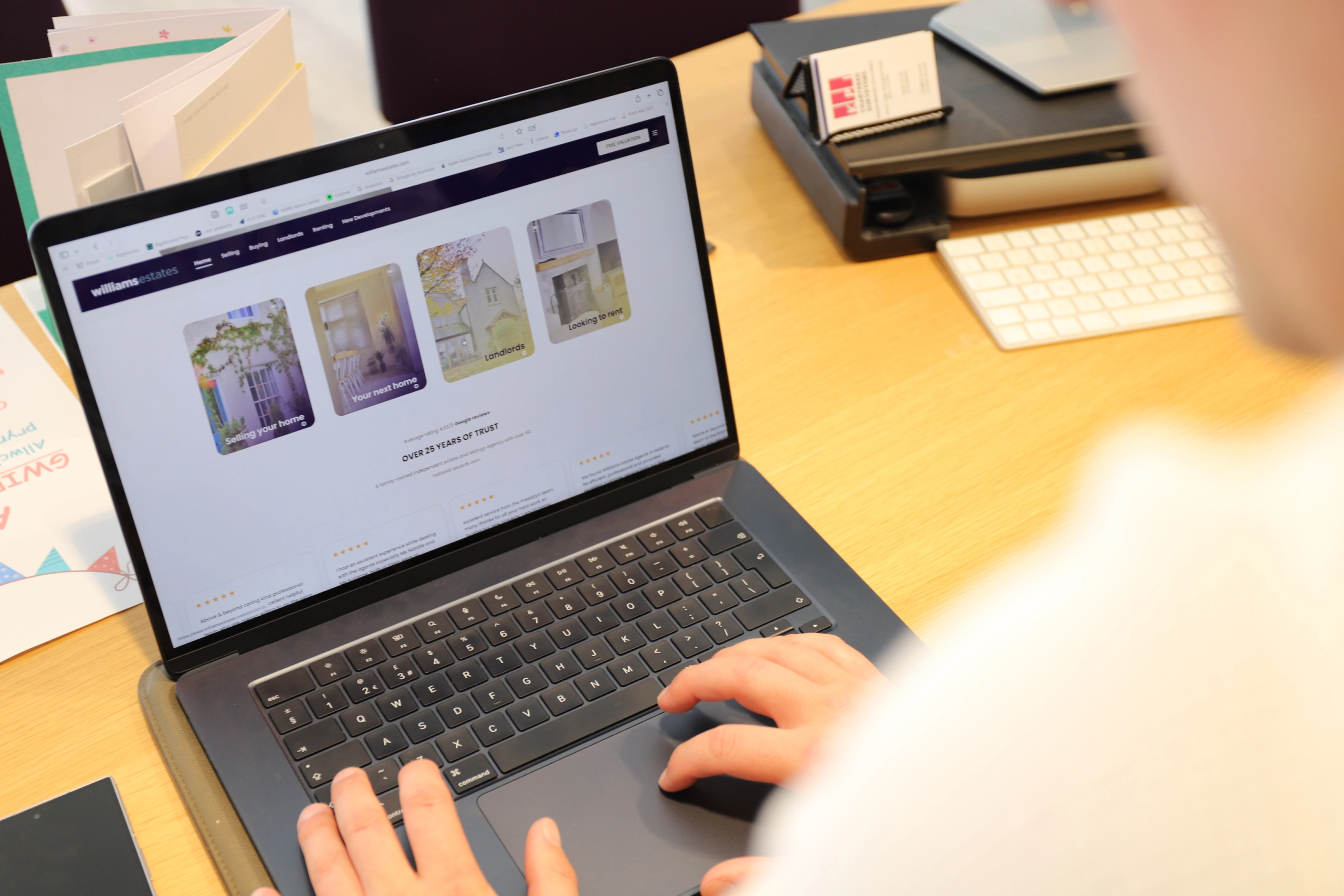Laptop displaying Williams estate agency website with property images, on a yellow desk with papers and office items