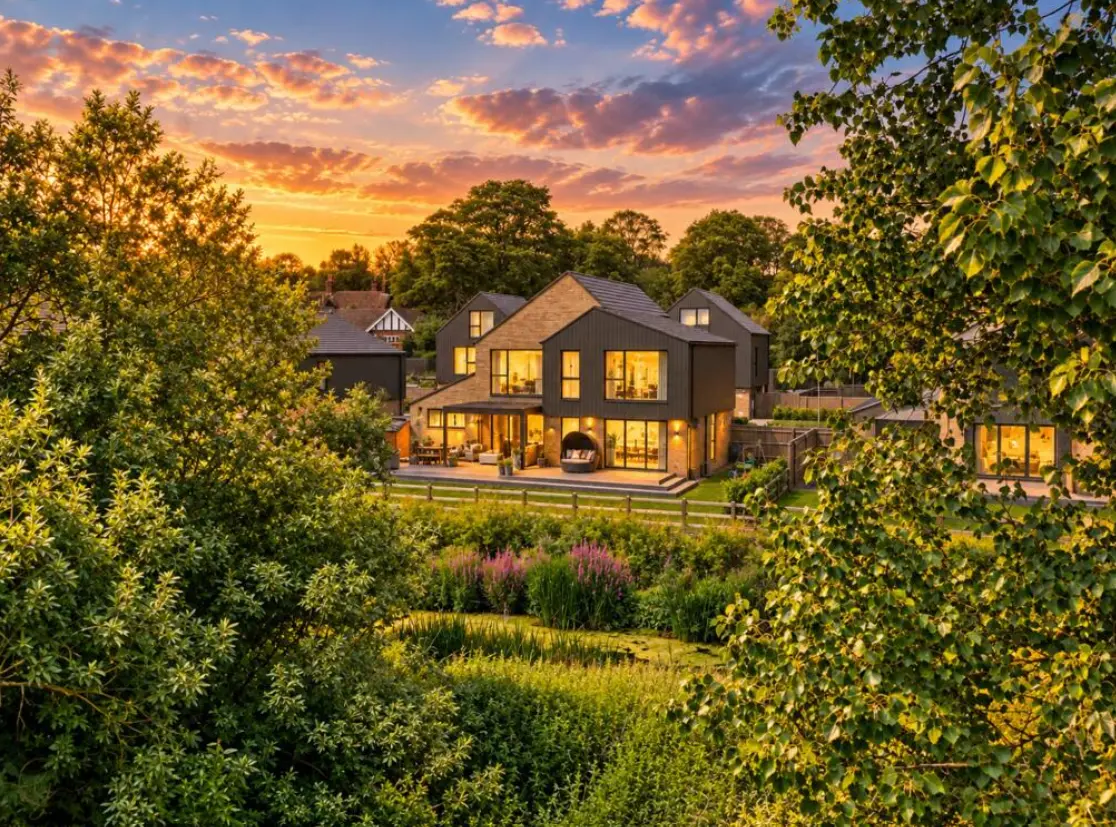 Modern two-storey house with dark exterior and illuminated windows at sunset, surrounded by mature trees and landscaped gardens with pink flowering shrubs