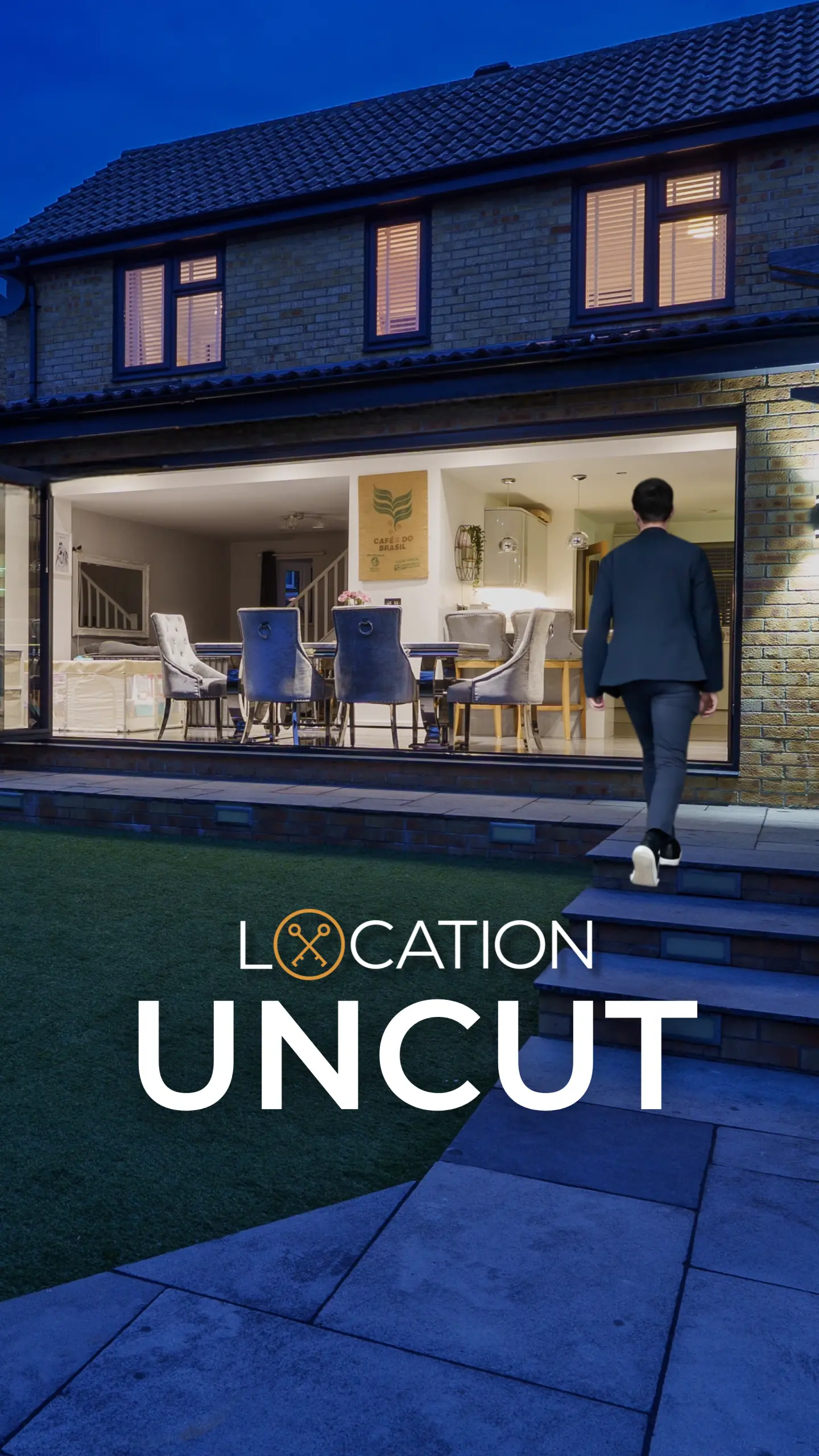 Modern two-storey brick house at dusk with illuminated interior showing dining area with blue chairs, person in dark suit walking on paved patio towards glass extension. Text overlay reads 'LOCATION UNCUT'