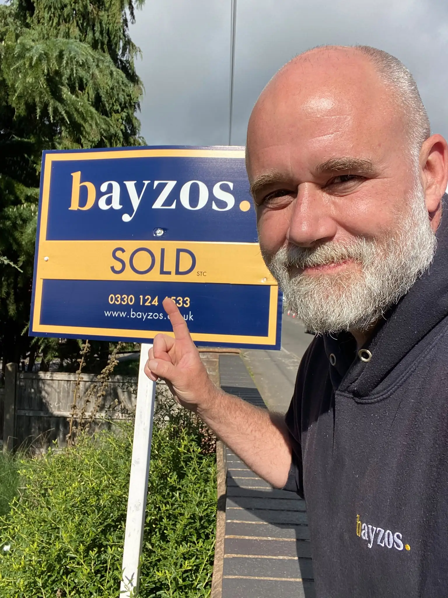 Man holding a Bayzos estate agency sold sign displaying contact number 0330 124 6533 and website www.bayzos.co.uk