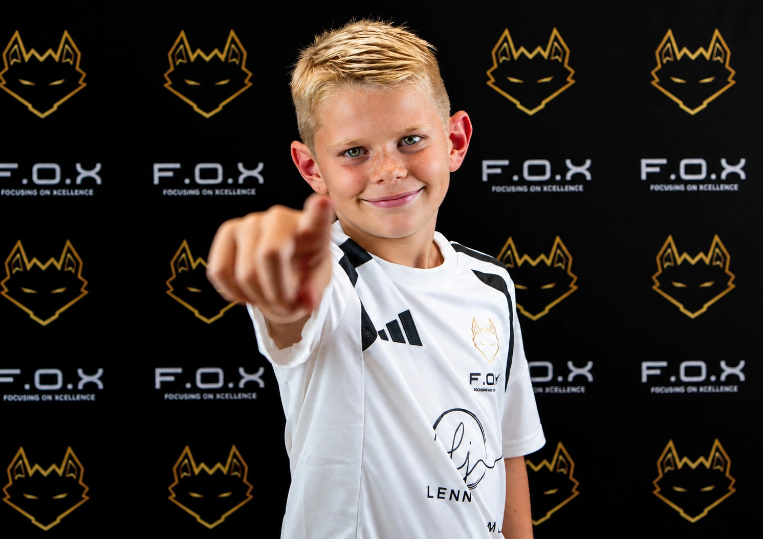 Child pointing at camera wearing white sports shirt with F.O.X branding and 'LENN' text, standing in front of black backdrop with gold fox head logos