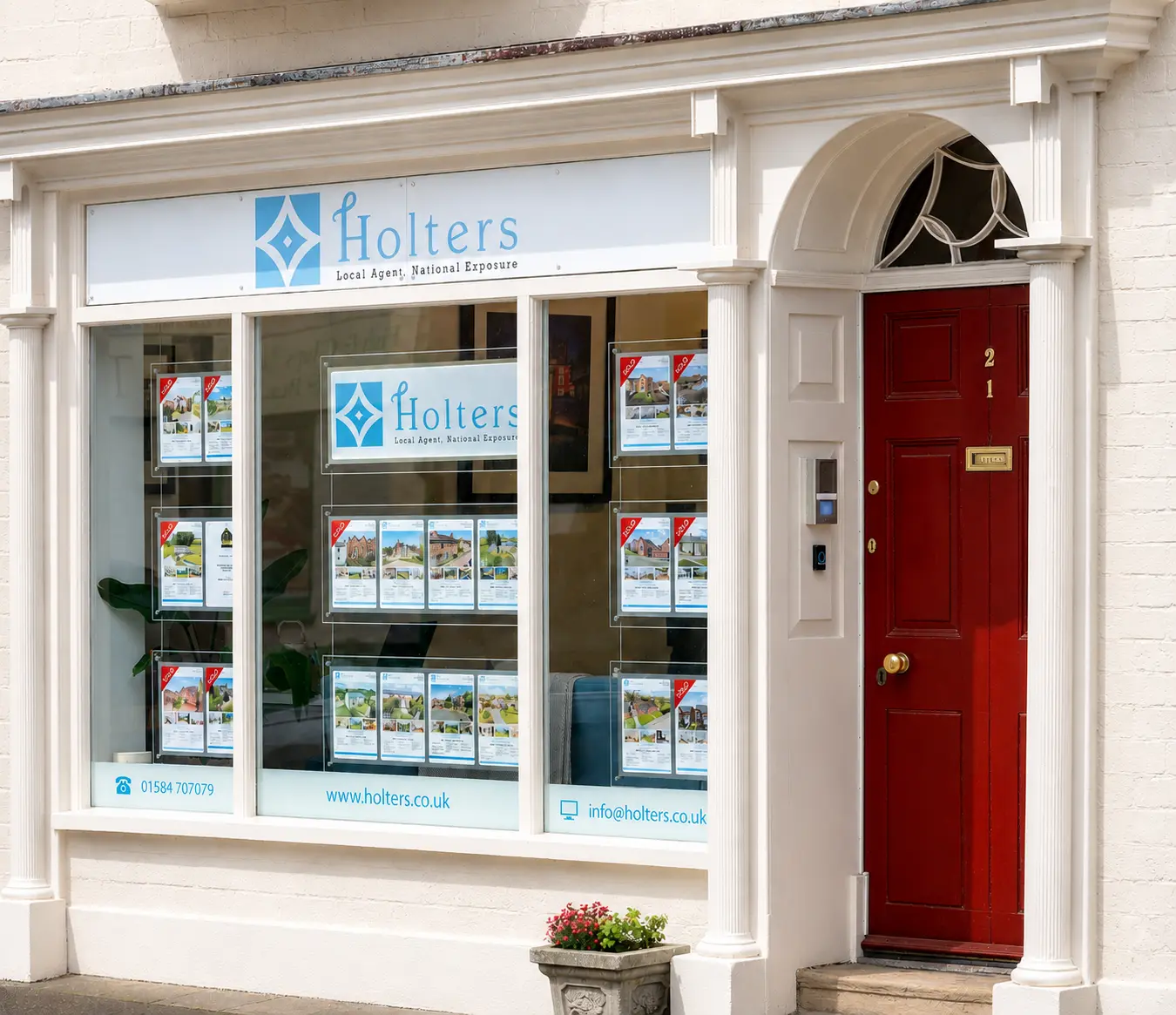 Holters estate agency storefront with white frame, blue logo, property listings displayed in windows, contact details 01584 707079 and www.holters.co.uk visible, red door numbered 21 to the right