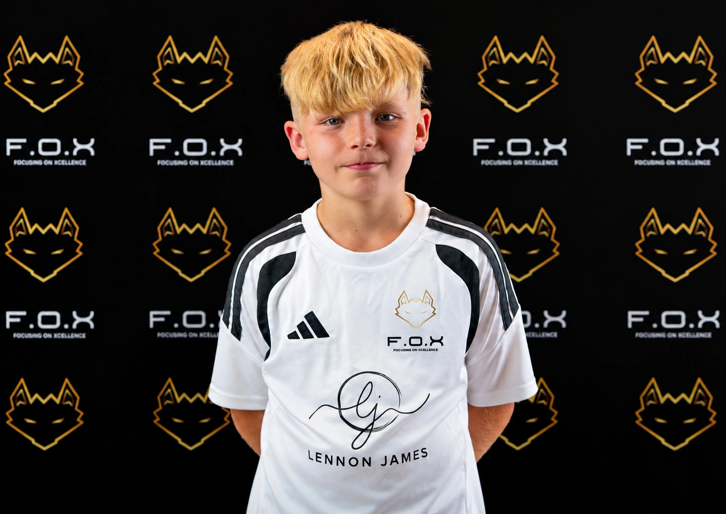 Young person wearing white sports jersey with F.O.X logo and 'Lennon James' printed on front, standing against black backdrop with repeating F.O.X branding
