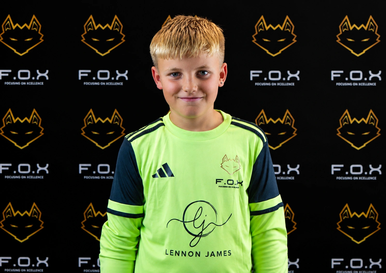 Young boy wearing a neon green F.O.X sports jersey with 'Lennon James' printed on it, standing in front of a repeating F.O.X logo backdrop