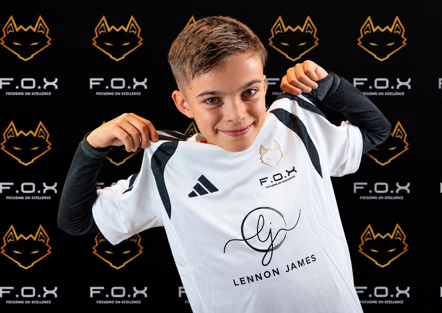 Young boy wearing white and black F.O.X sports jersey with signature and name 'Lennon James' printed on it, posing in front of branded backdrop