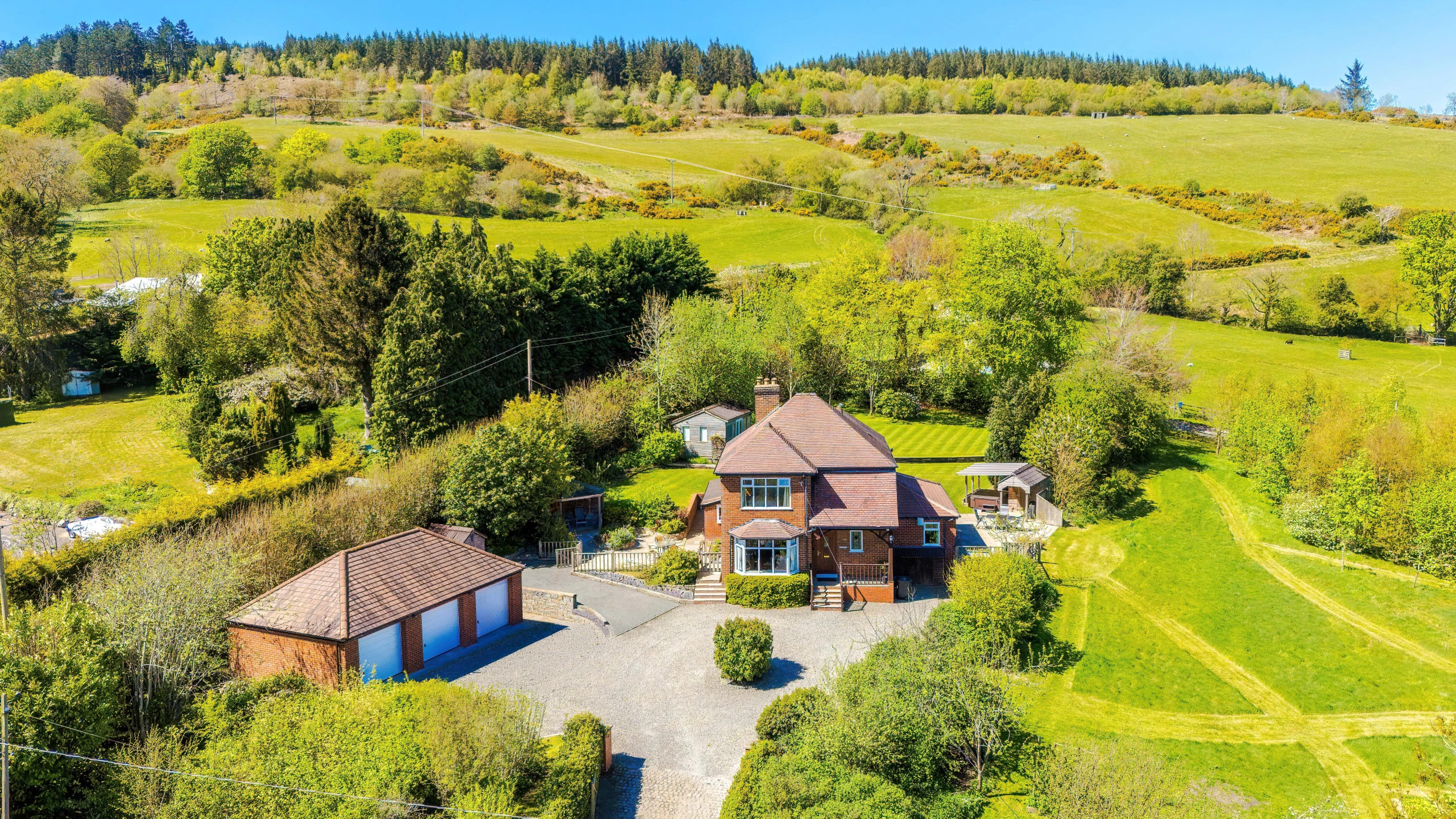 Aerial view of a brick house with pitched roof and driveway, surrounded by green fields, trees, and a detached garage with blue doors