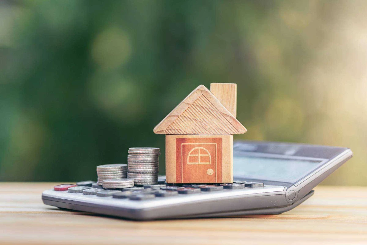 Wooden house model with stacked coins on a calculator, blurred green background
