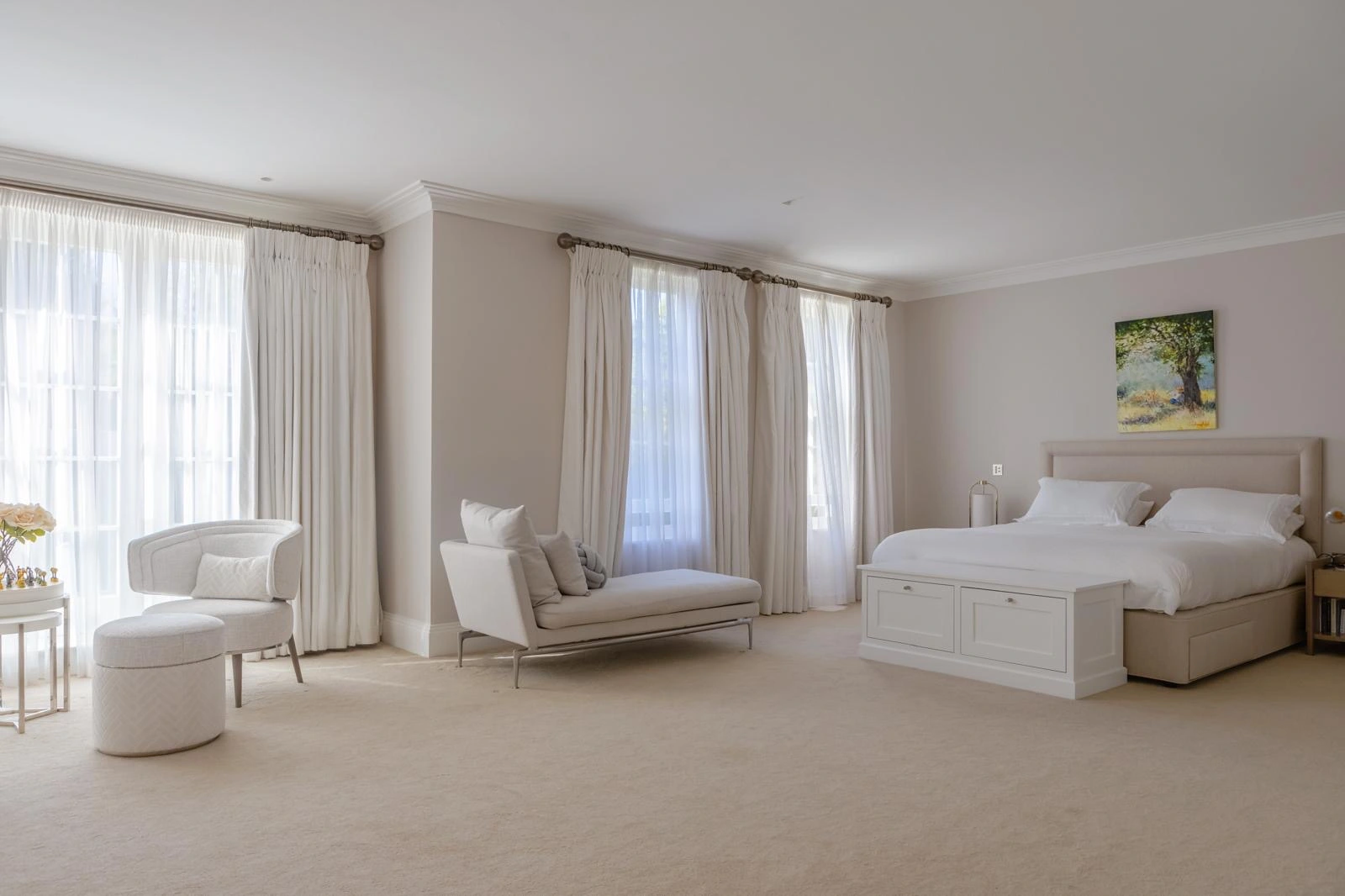 Spacious bedroom with neutral décor, featuring a bed with upholstered headboard, cream armchair, daybed with cushions, multiple windows with floor-to-ceiling curtains, and landscape artwork on the wall