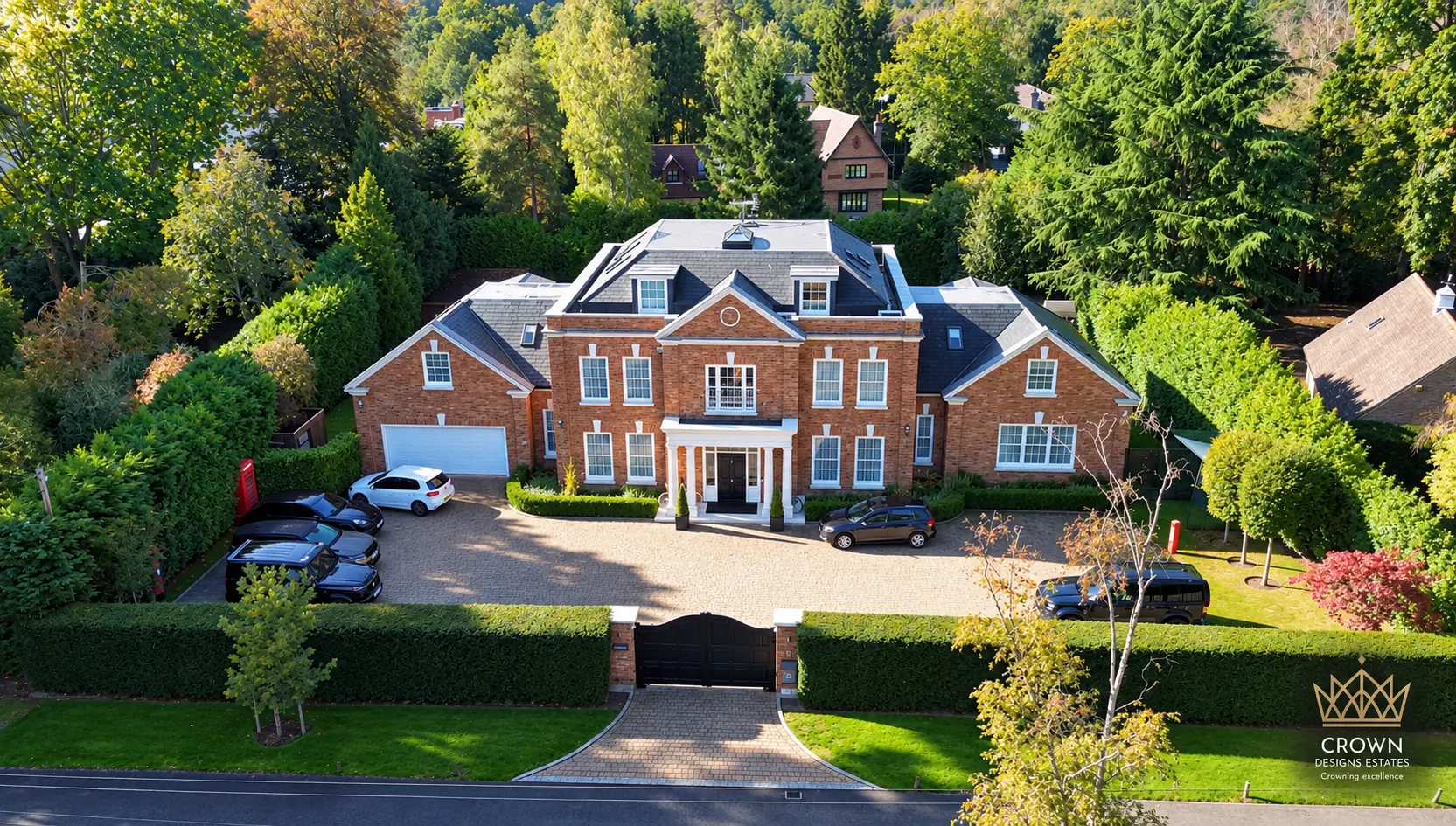 Aerial view of a large brick detached house with slate roof, white-trimmed windows, columned entrance portico, and driveway with multiple parked cars, surrounded by mature trees and hedging