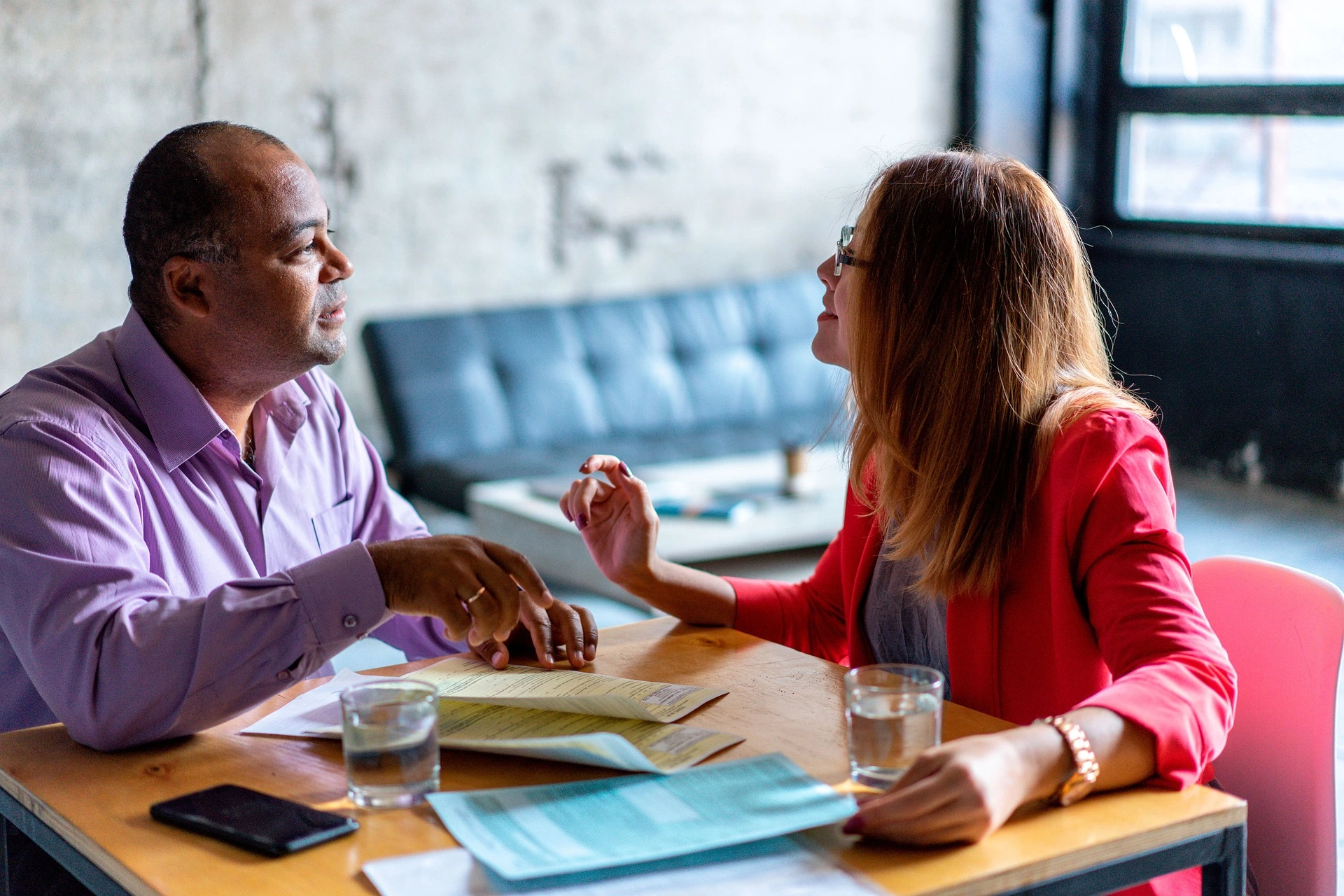 Two people sitting at a wooden table in conversation, with papers and glasses of water in front of them
