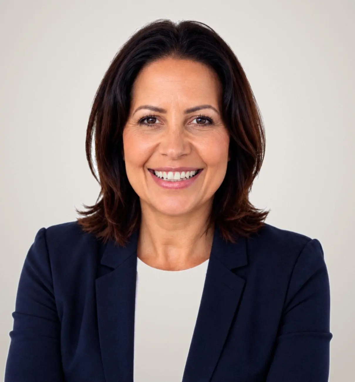 Professional headshot of a woman with dark brown hair wearing a dark navy blazer over a white top, smiling at the camera against a neutral grey background