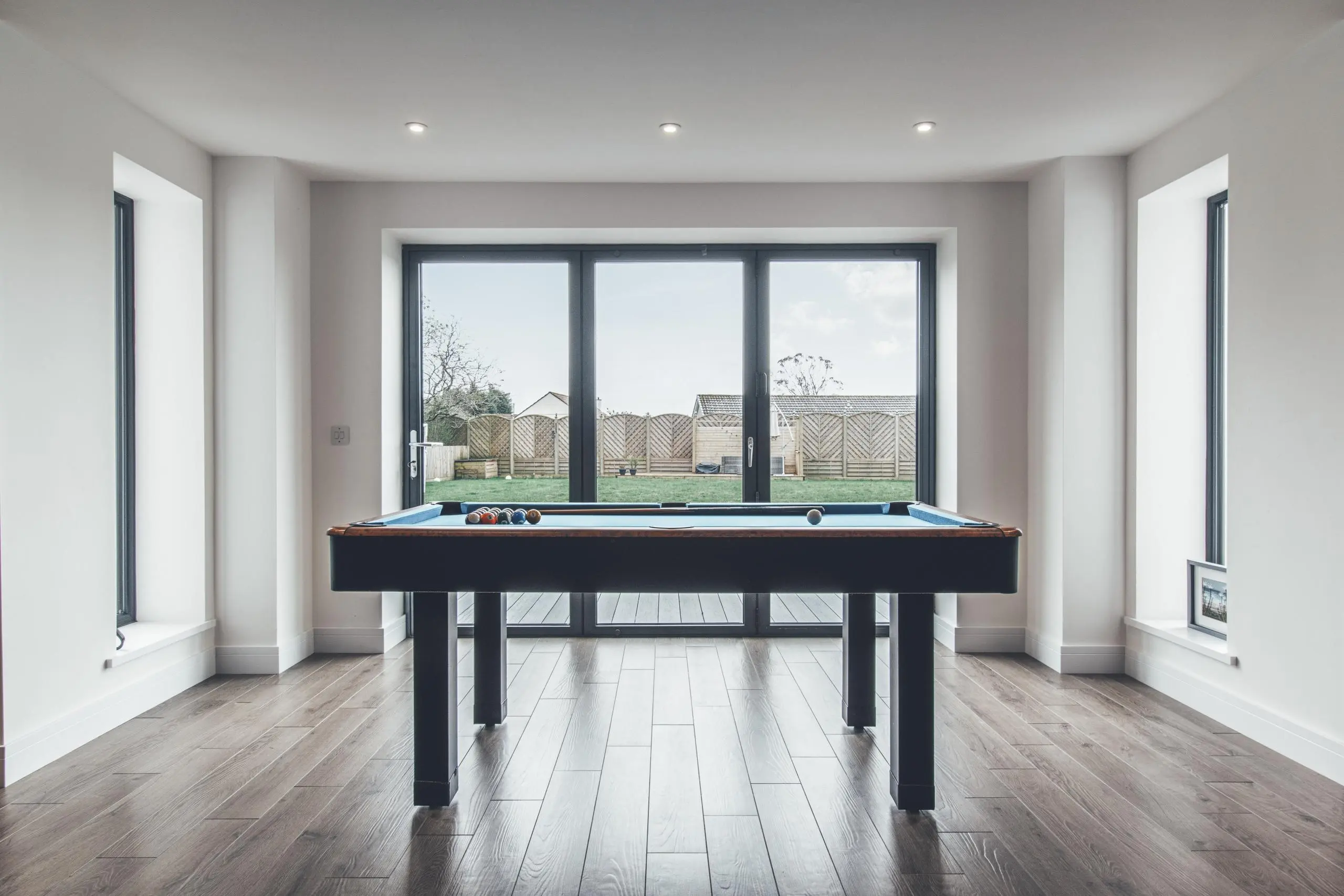 Modern room with pool table, wooden flooring, and large windows overlooking garden with fencing