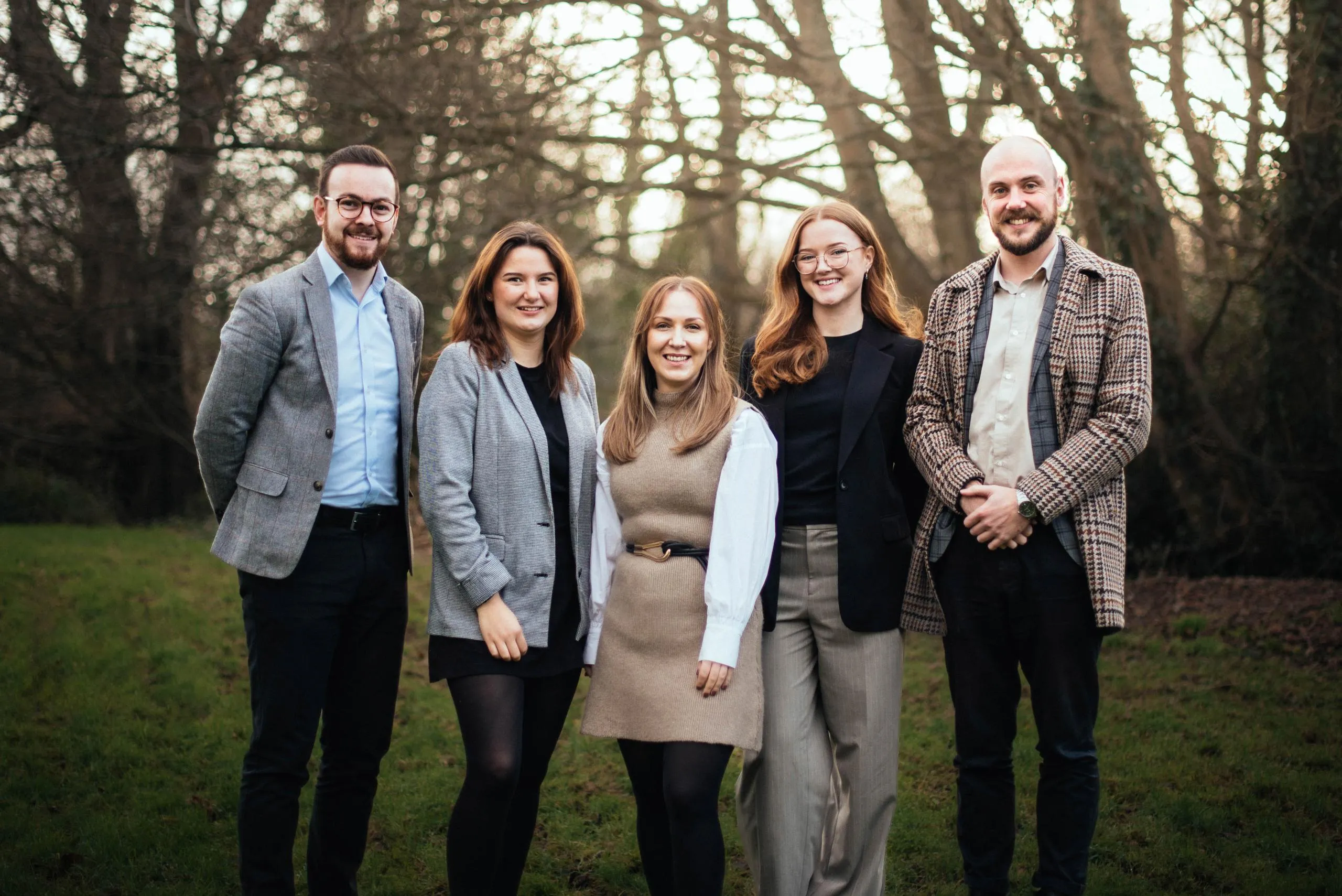 Five people standing in a line outdoors in front of bare trees, wearing business casual clothing