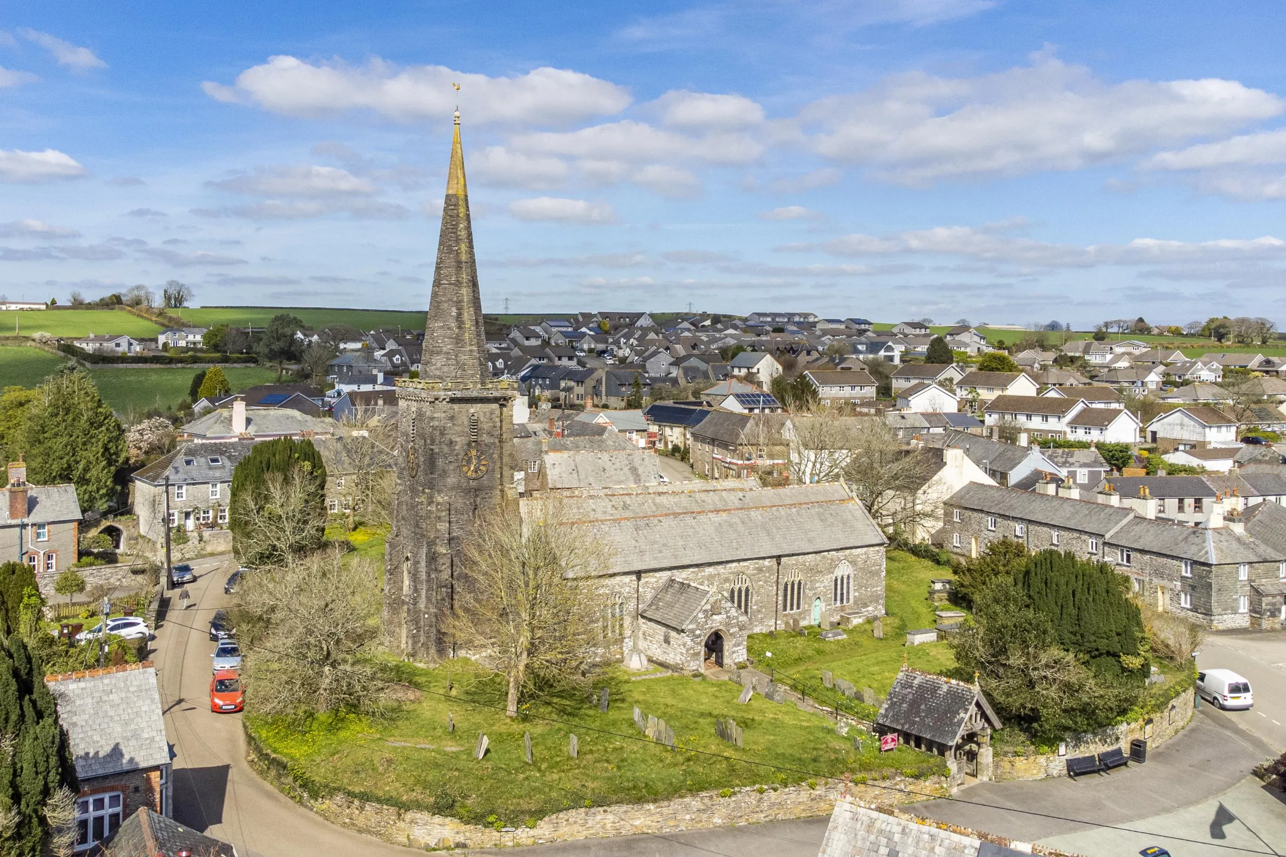 Stone church with tall spire in village centre, surrounded by residential houses and green fields