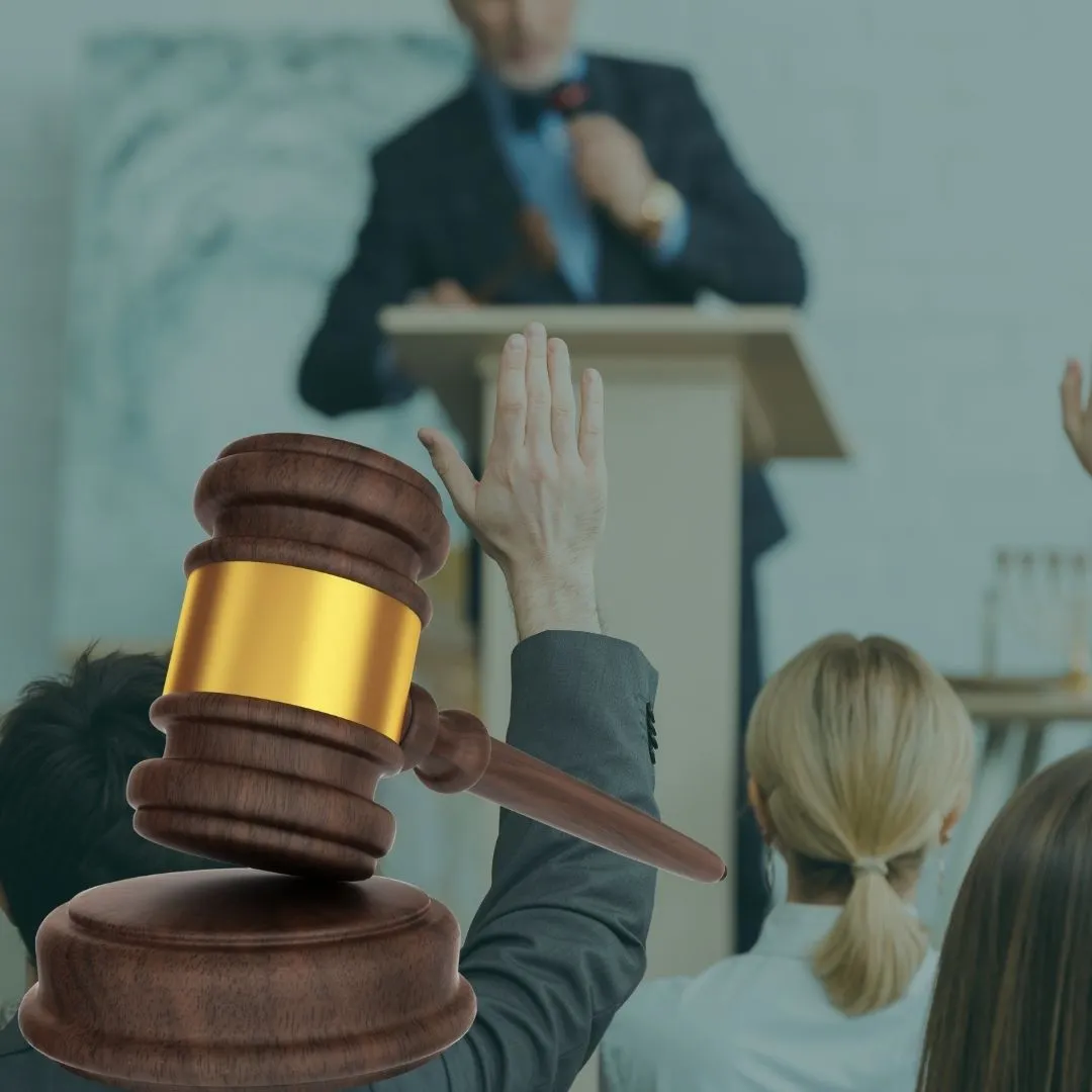 Hand holding a gavel with gold band, raised in front of a person at a podium and audience members