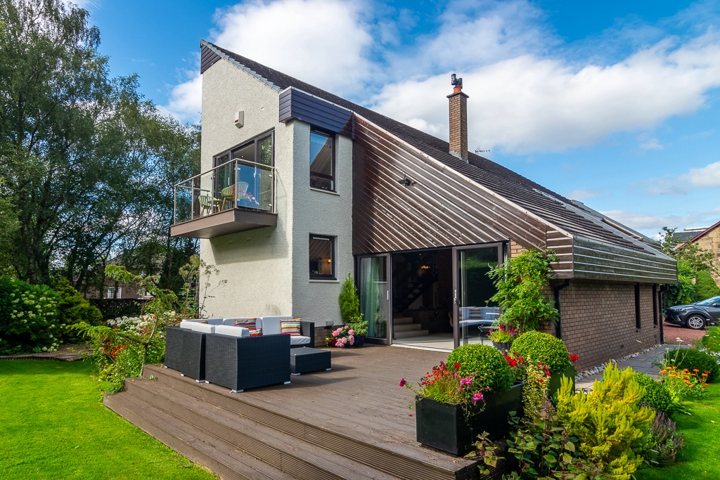 Modern house with white and dark rendered exterior, metal roof, balcony, patio with outdoor seating, and landscaped garden