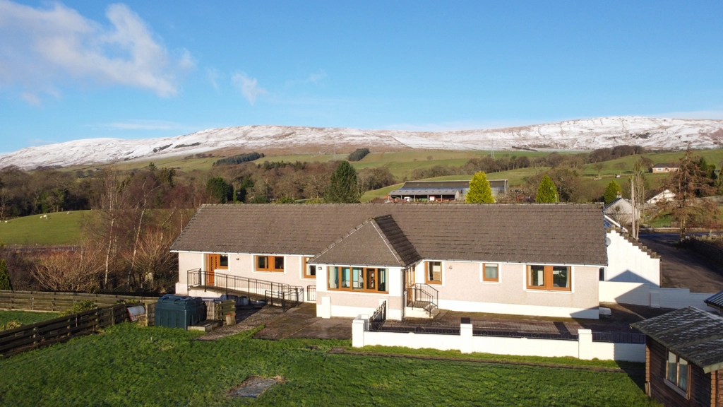 Modern single-storey white house with grey roof and wooden windows, set in rural landscape with snow-capped mountains in background