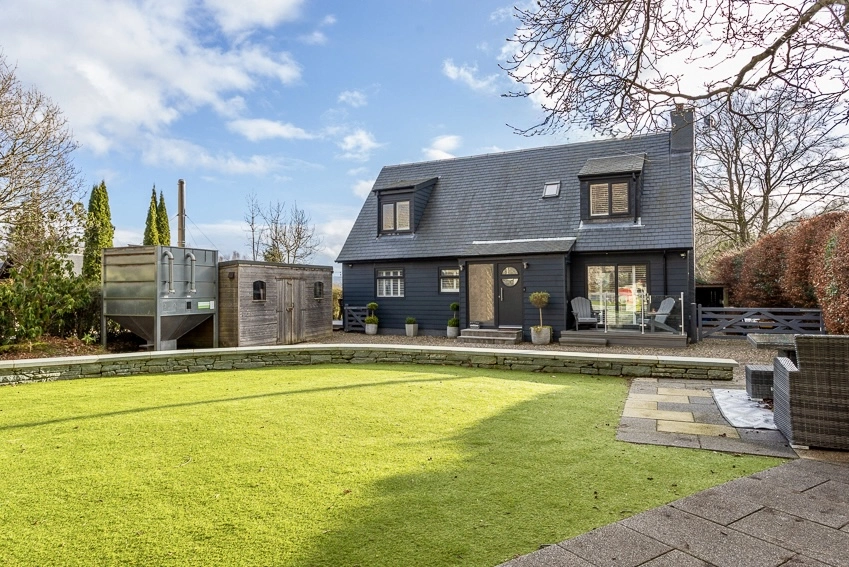 Modern dark-clad house with pitched roof and dormer windows, viewed from garden with lawn and paved terrace