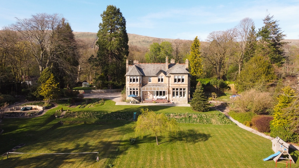 Large stone country house with manicured gardens, mature trees and rolling hills in background