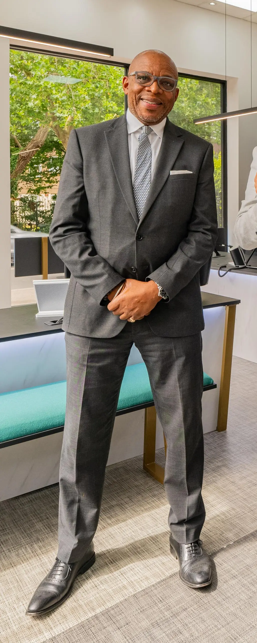 Man in grey suit and glasses standing in modern office with large window overlooking garden