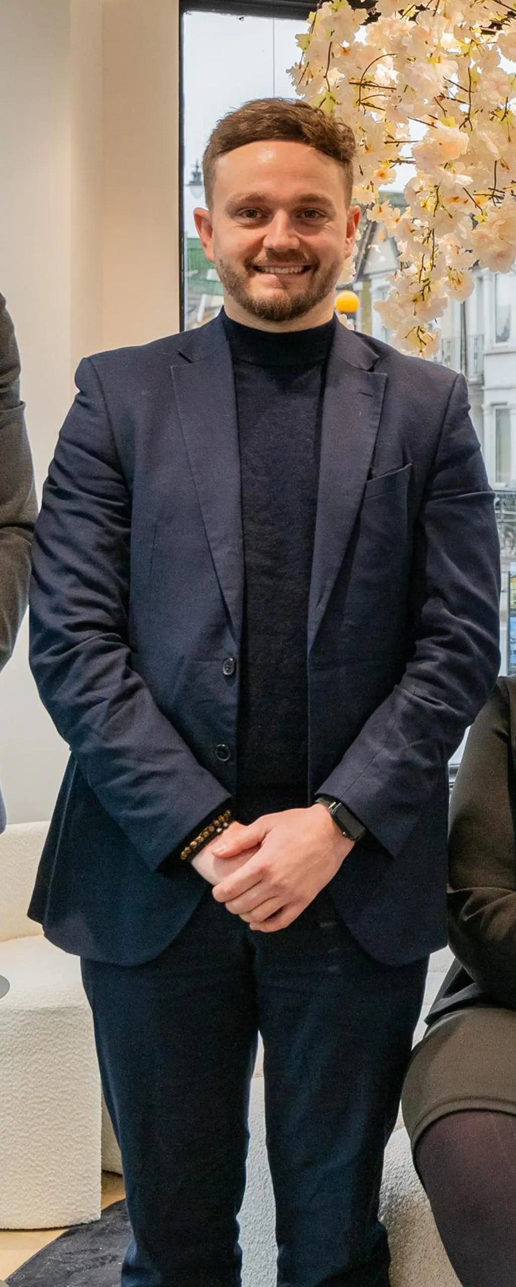 Man in dark navy blazer and black turtleneck standing indoors by a window, with dried white flowers visible above