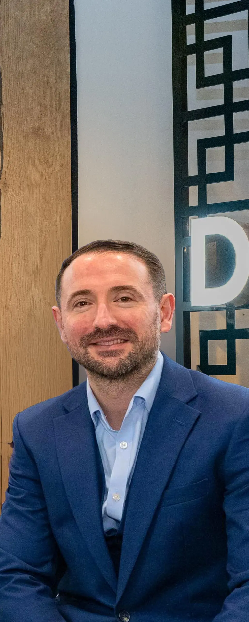 Man in navy blazer and light blue shirt, smiling at camera, standing in front of wooden panelling and geometric patterned screen