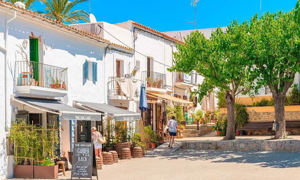 Mediterranean-style white buildings with terracotta roofs, palm trees, and a café with outdoor seating in a sunny plaza