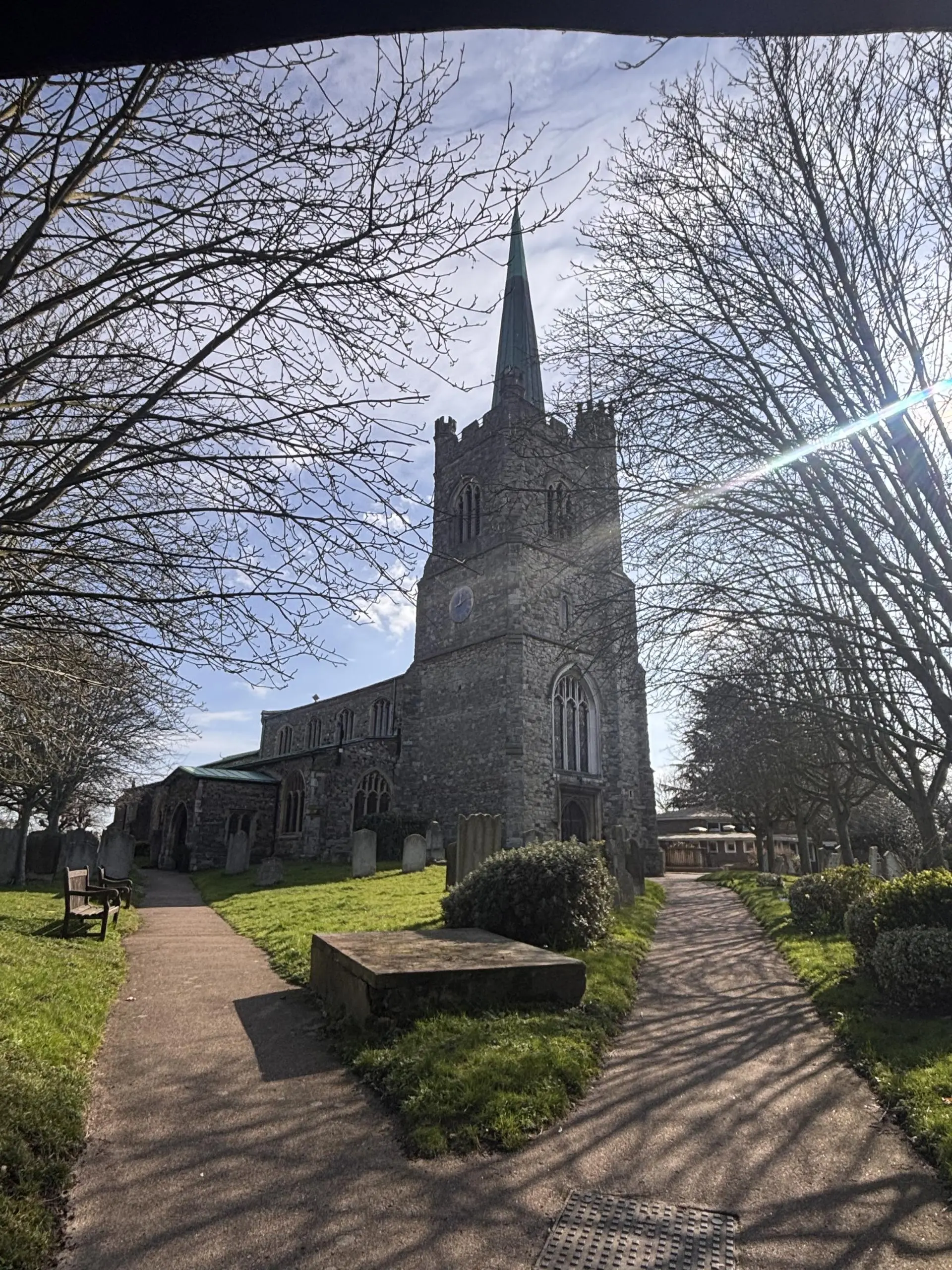 Stone church with tall spire, surrounded by bare trees and gravestones, with pathways and green grass in foreground