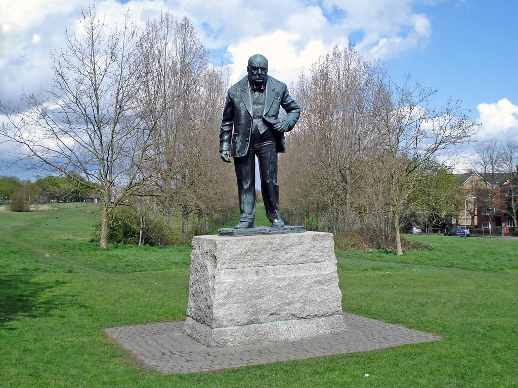 Bronze statue of a man in a suit standing on a stone plinth in a park with bare trees and grass