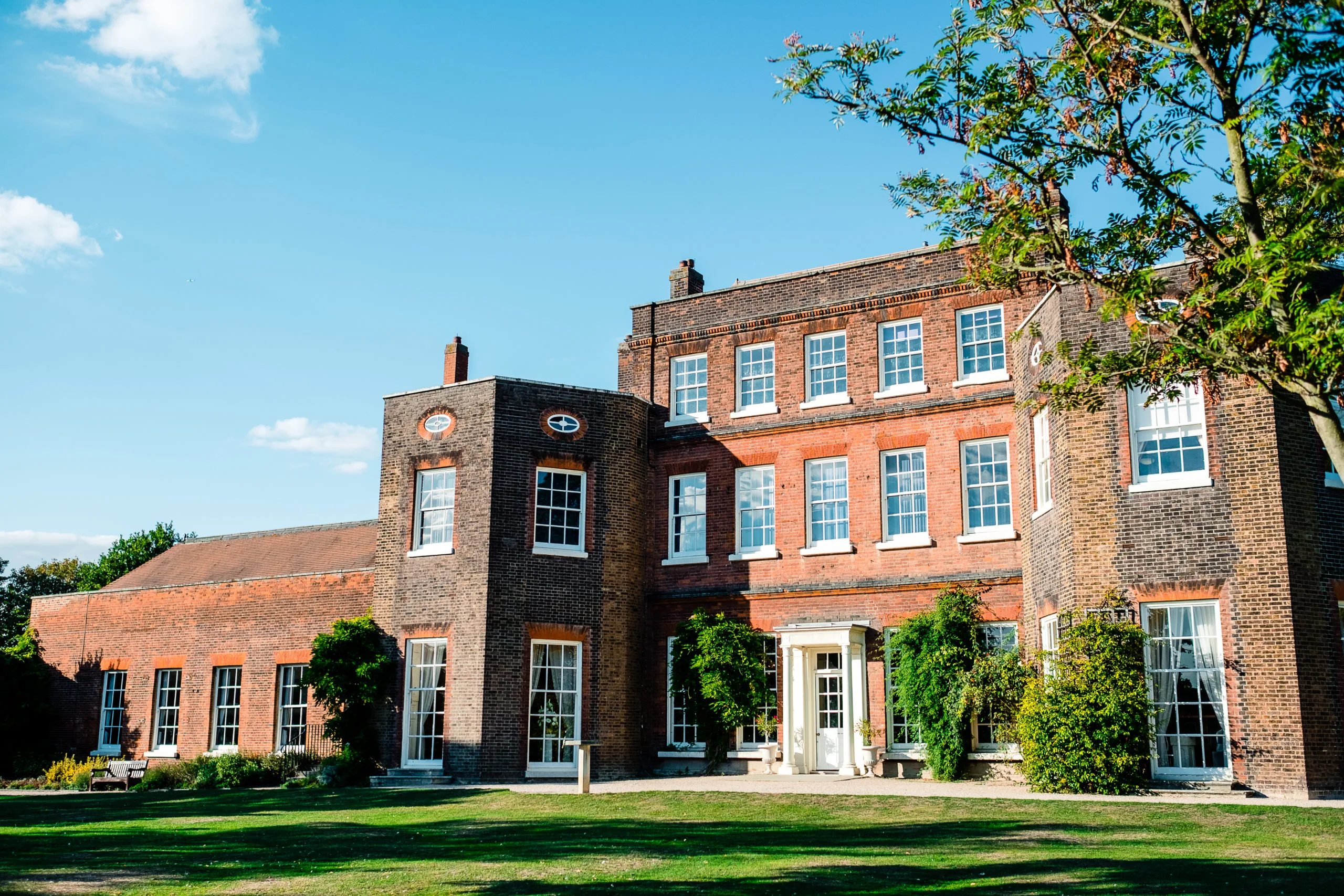 Large brick Georgian house with ivy-covered entrance, manicured lawn, and clear blue sky