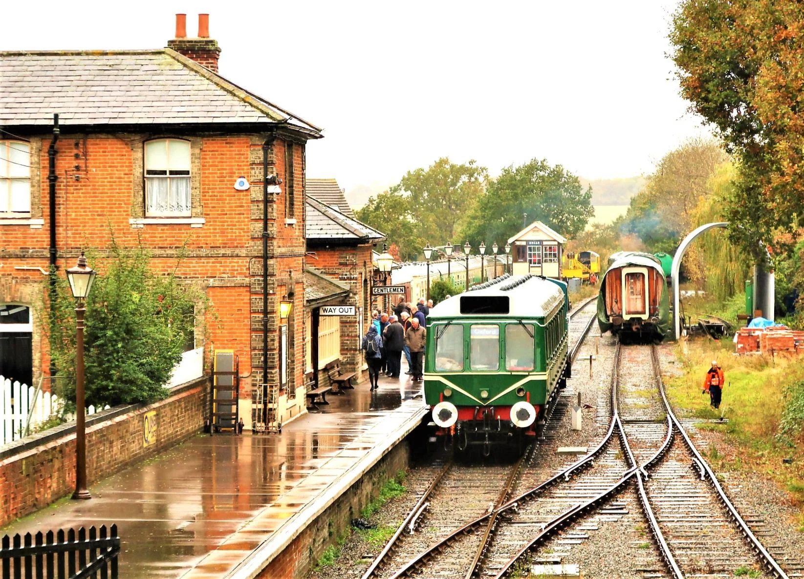 Red brick Victorian station building with green heritage railway train at platform, passengers boarding, autumn countryside beyond