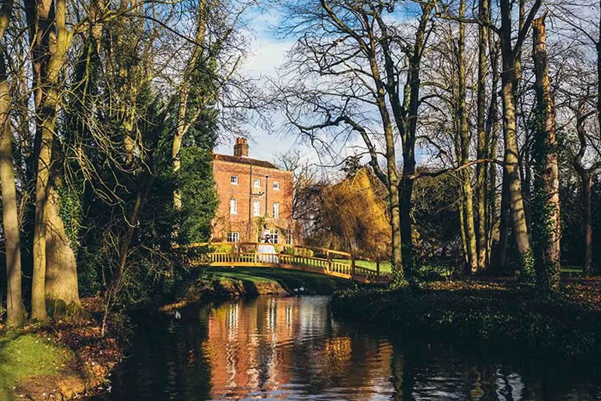 Red brick building with chimneys viewed across a stream and tree-lined grounds, with a yellow footbridge in the foreground