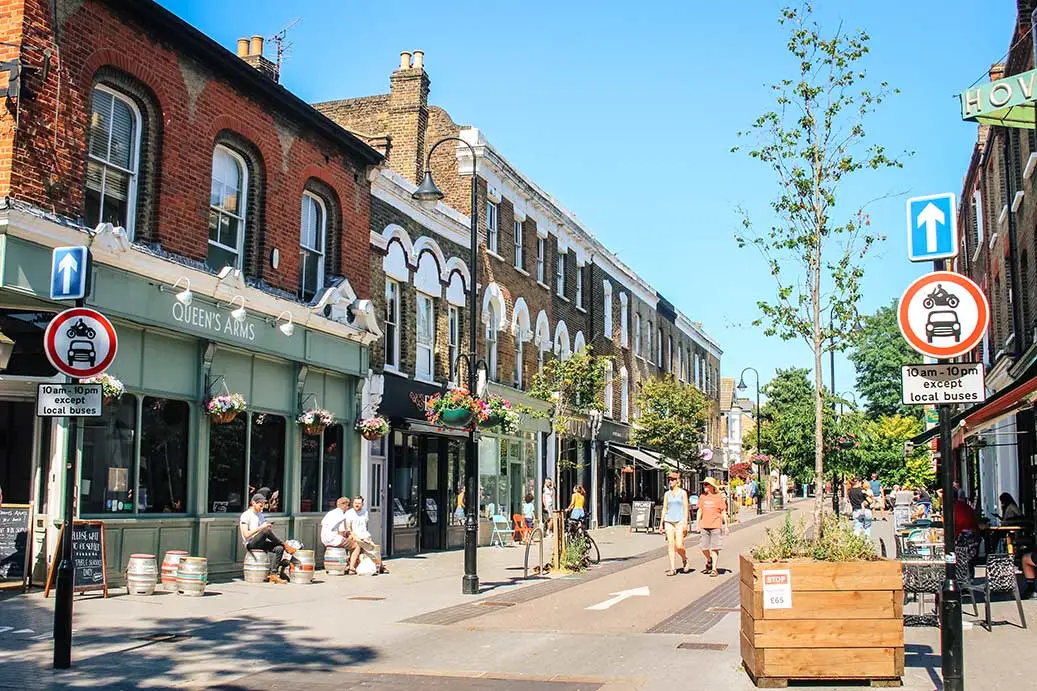 Walthamstow Village high street with Victorian brick buildings, shops including Queen's Arms pub, pedestrians, trees and traffic signs on a sunny day