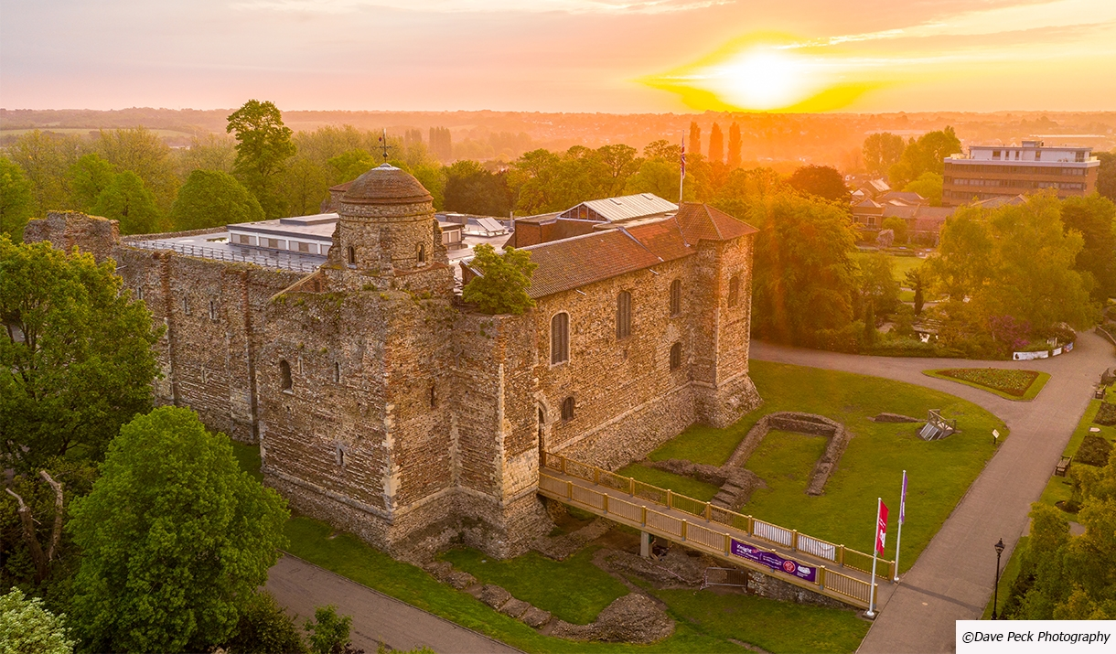 Stone castle with circular tower at sunset, surrounded by green trees and manicured grounds with visitor facilities visible