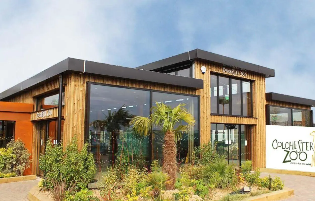 Modern building with wooden cladding and dark roof, large windows, palm tree and landscaping, Colchester Zoo sign visible