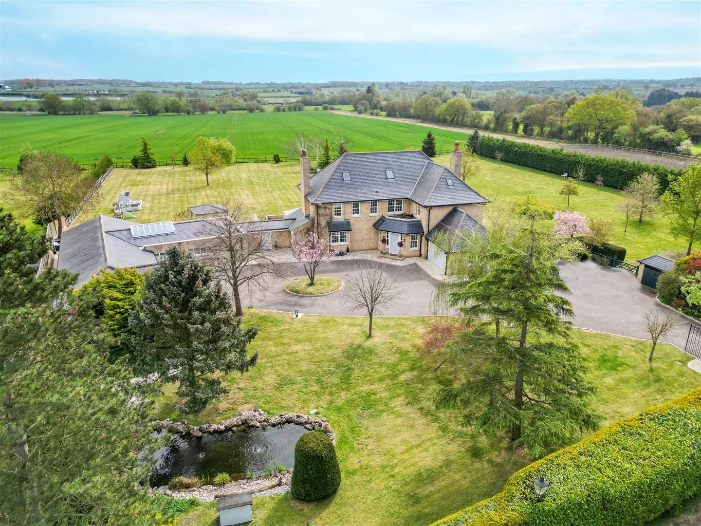 Aerial view of a large brick country house with slate roof, surrounded by manicured gardens, trees, and open countryside with fields beyond