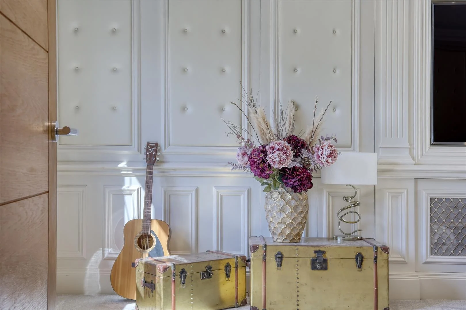 Hallway with white panelled walls, acoustic guitar, yellow vintage suitcases, and white vase with pink and burgundy flowers and dried grasses