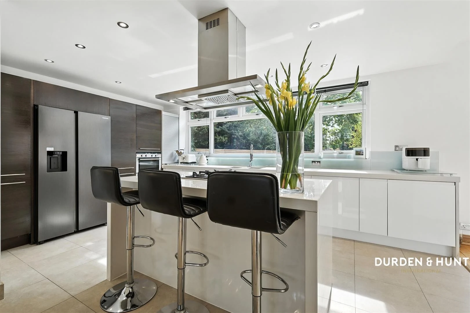 Modern kitchen with white and dark grey cabinetry, island with black bar stools, stainless steel appliances, and large windows overlooking greenery