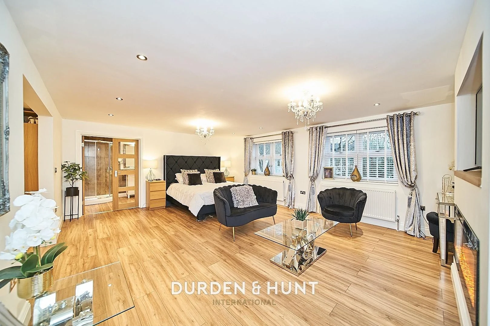 Spacious bedroom with wooden flooring, double bed with black headboard, black armchair, ensuite bathroom, large windows with shutters and curtains, crystal chandeliers, and wood-grain doors