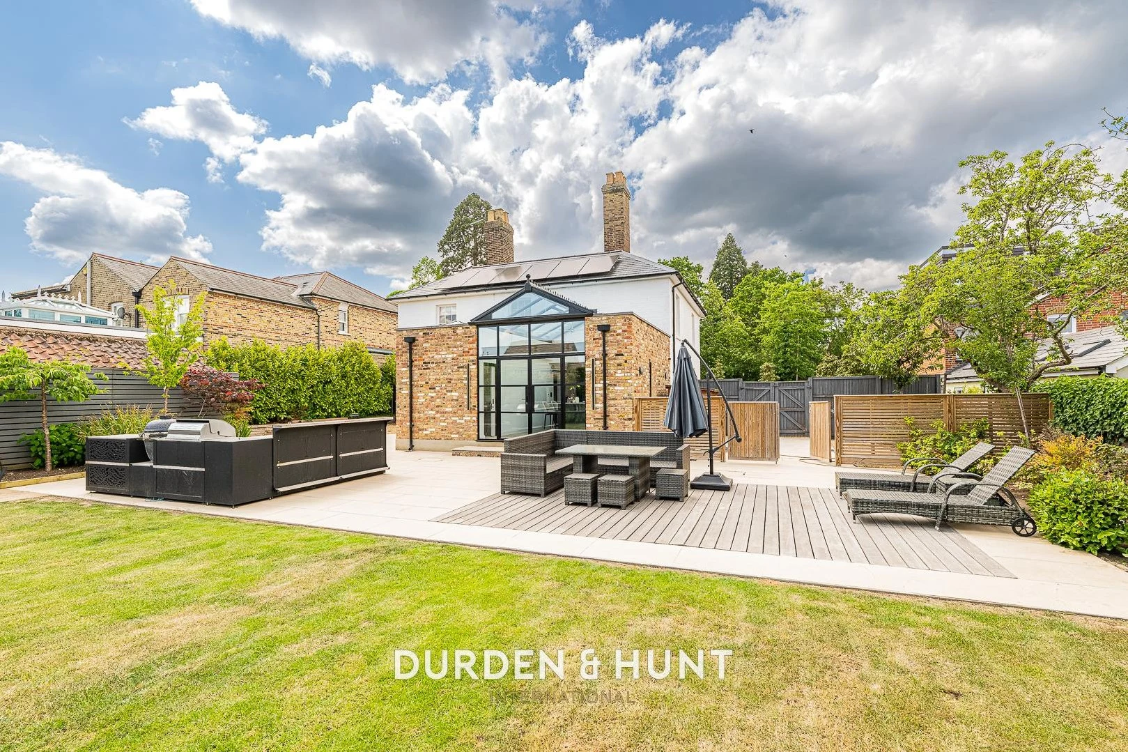 Modern brick extension with glass roof, patio seating area, wooden decking, and manicured garden with fencing