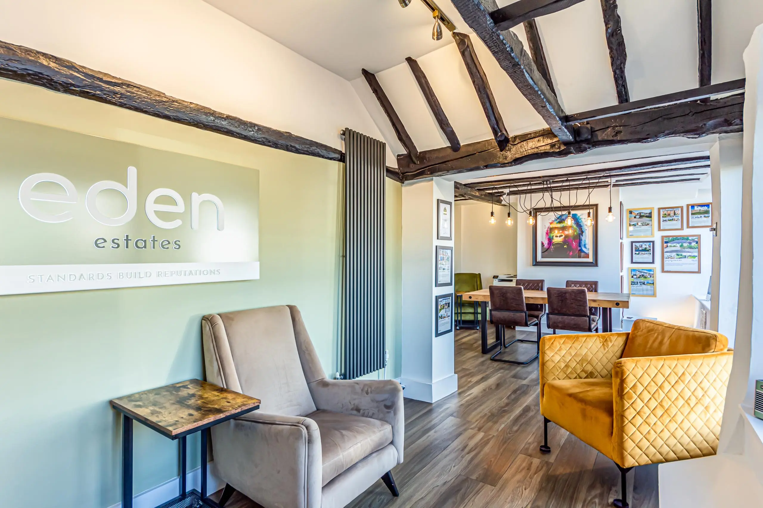 Eden Estates office interior with exposed wooden beams, green accent wall with white logo, grey armchair, mustard yellow leather chair, and meeting area with framed property photographs