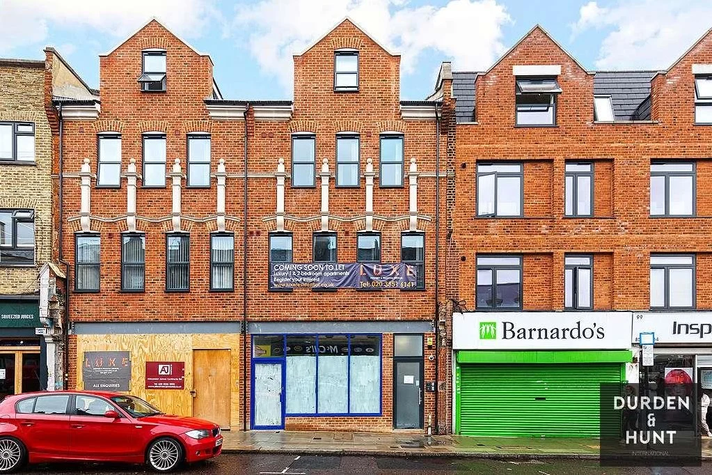 Row of Victorian red brick buildings with boarded-up ground floor shops, including Barnardo's and Durden & Hunt storefronts, with a red car parked outside