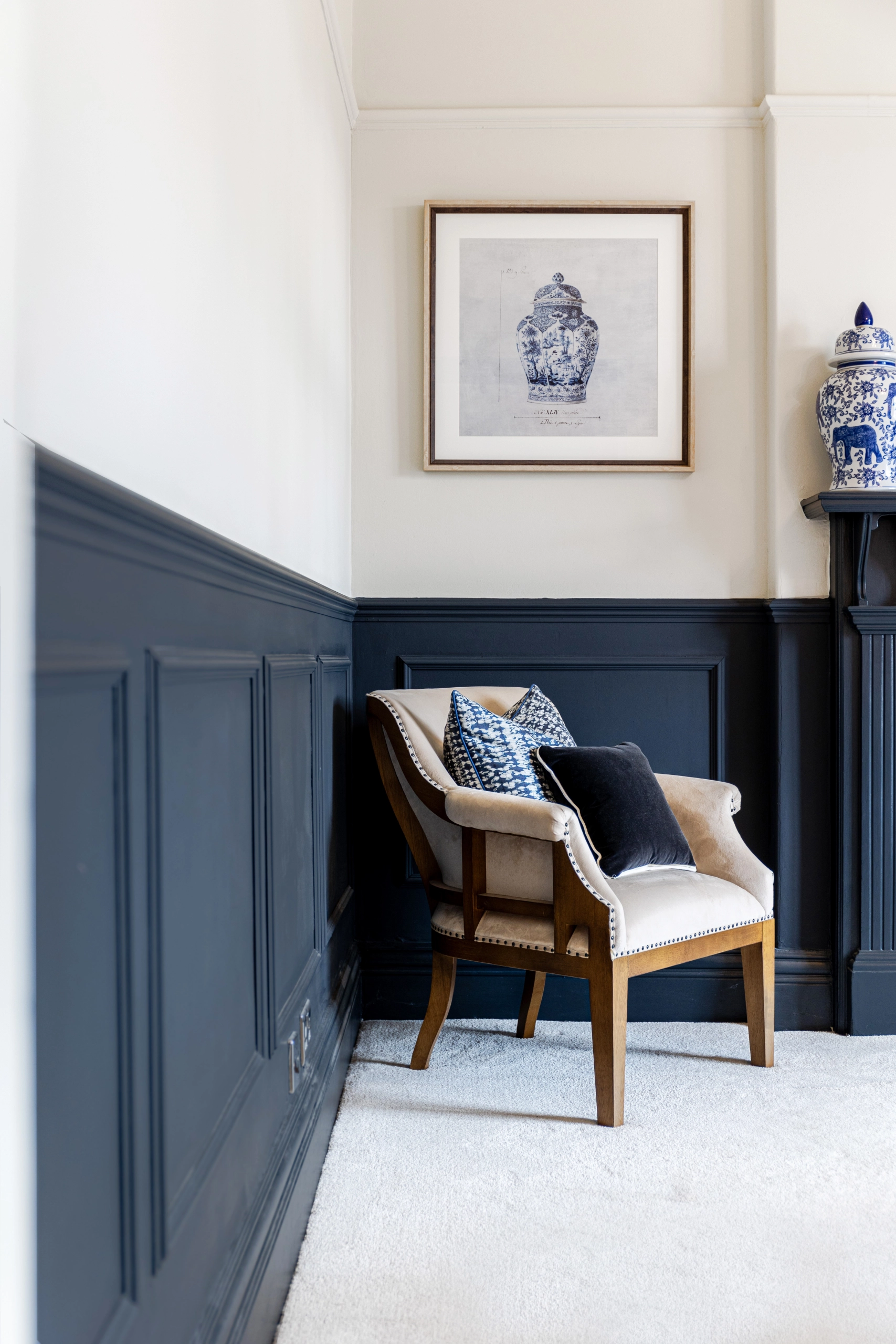 Interior hallway with dark blue panelled walls, cream upper walls, upholstered armchair with wooden frame and blue patterned cushions, framed blue and white ginger jar print above, and decorative blue and white porcelain jar on pedestal