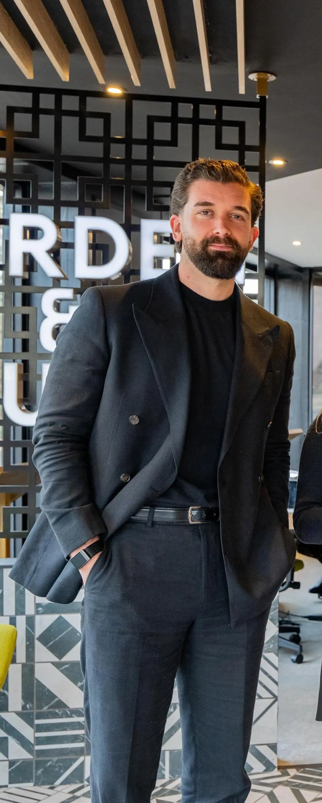 Man in dark double-breasted blazer and black shirt standing in modern office interior with geometric wall pattern and partial "RDE" signage visible