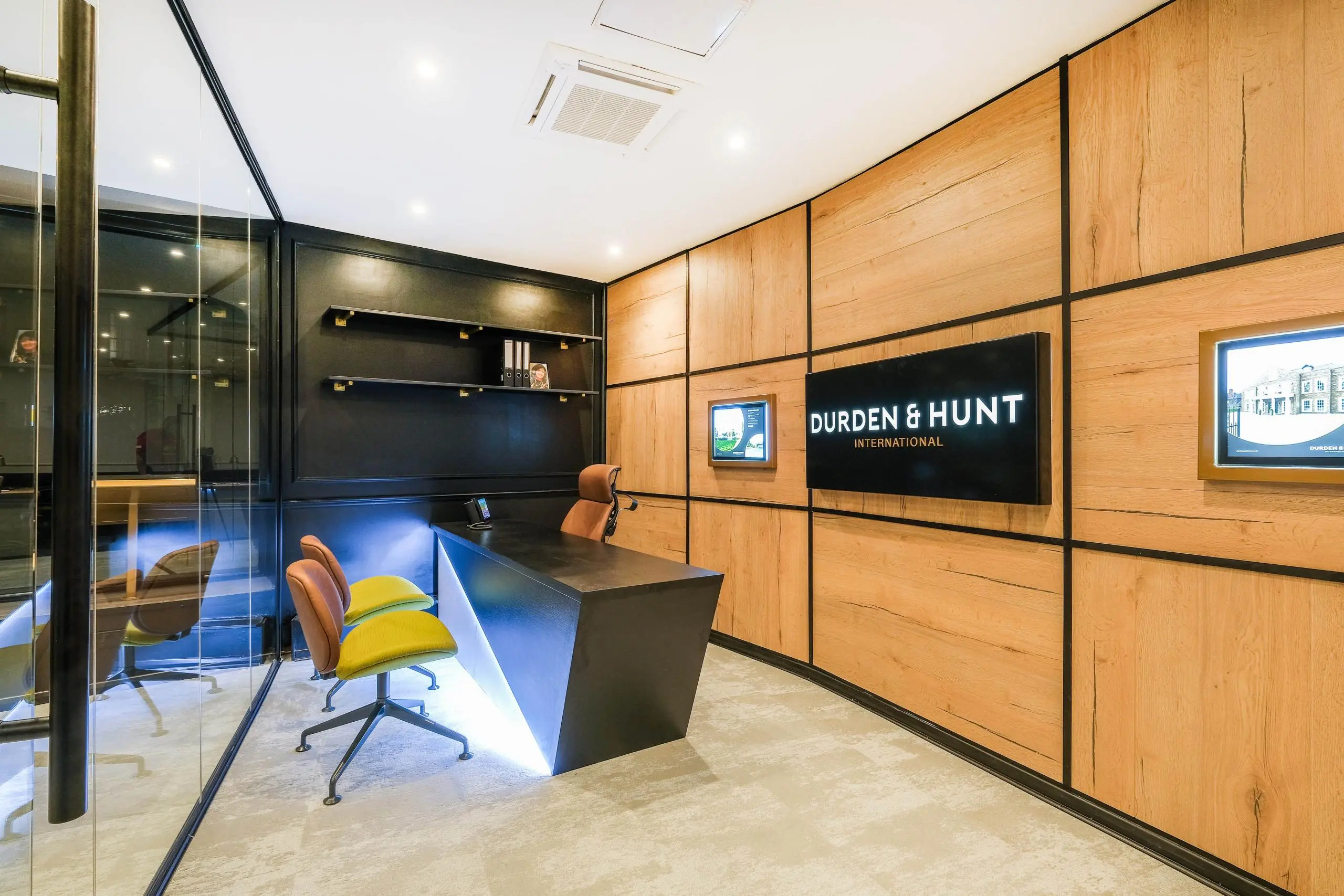 Modern office interior with wooden wall panelling, blue desk, orange office chairs with yellow cushions, glass partition, and "Durden & Hunt International" sign on wall