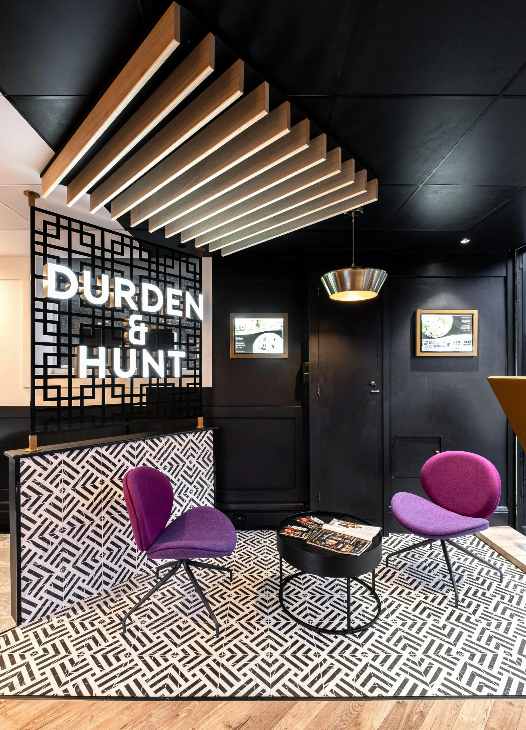 Durden & Hunt estate agency office reception with geometric patterned flooring, purple chairs, illuminated wooden ceiling beams, and black lattice screen with white lettering