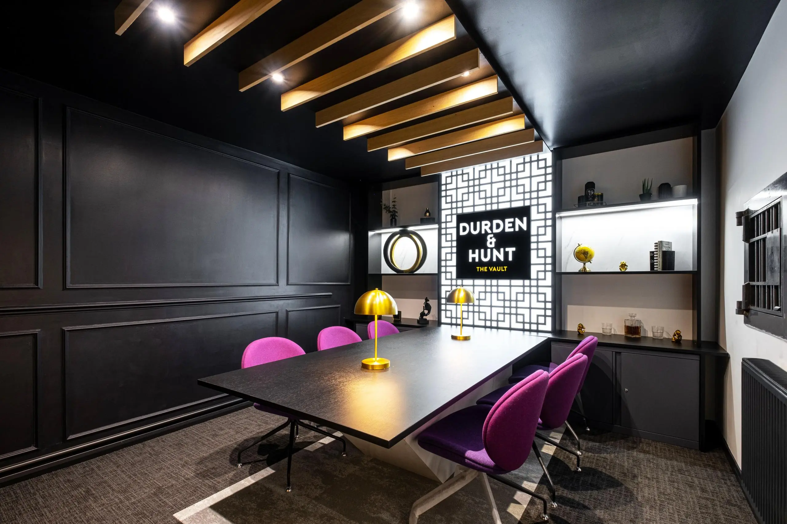 Modern office meeting room with black walls, wooden ceiling beams with integrated lighting, a dark table with pink chairs, and a "Durden & Hunt The Vault" branded display wall with shelving