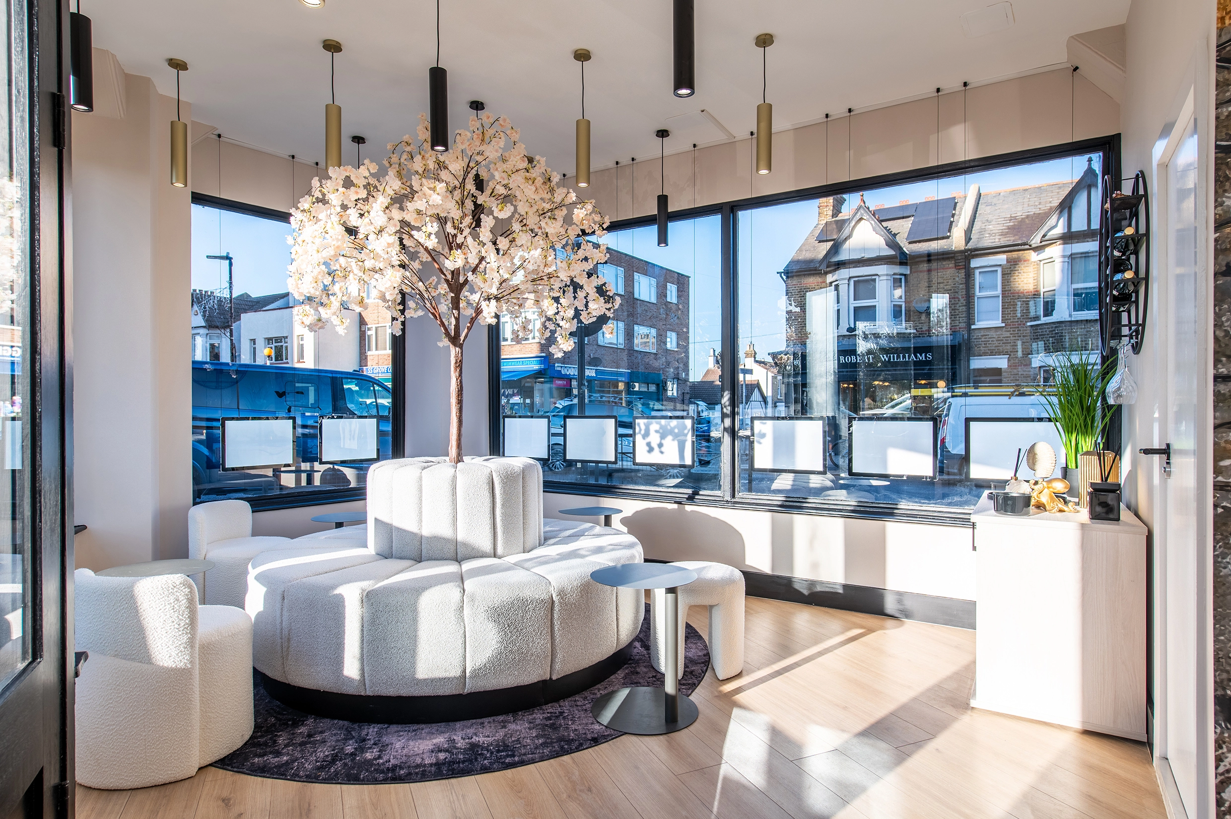 Modern estate agency office interior with curved white seating, decorative tree, pendant lights, and street-facing windows overlooking Victorian buildings